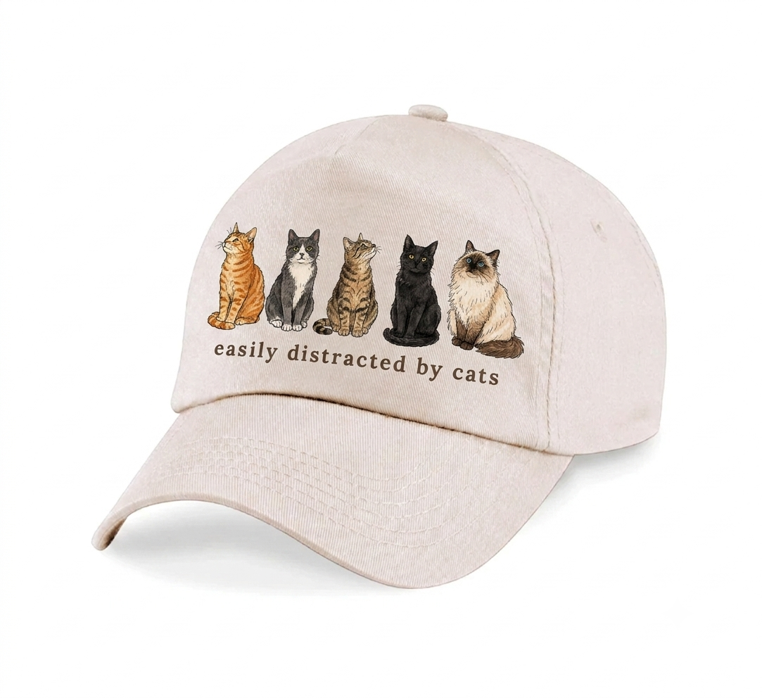 A light beige baseball cap featuring a graphic of five different cats sitting in a row above the text 'easily distracted by cats'.