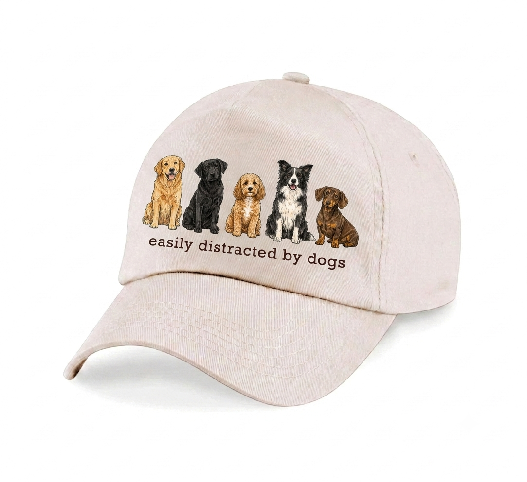 A light beige baseball cap featuring an embroidered graphic of five different dog breeds and the text 'easily distracted by dogs' on the front panel, set against a plain white background.