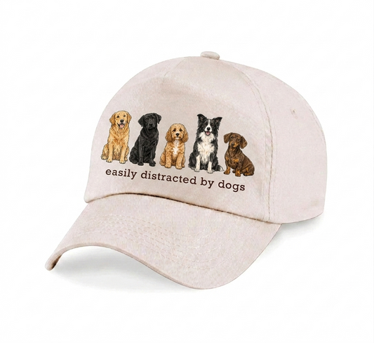 A light beige baseball cap featuring an embroidered graphic of five different dog breeds and the text 'easily distracted by dogs' on the front panel, set against a plain white background.