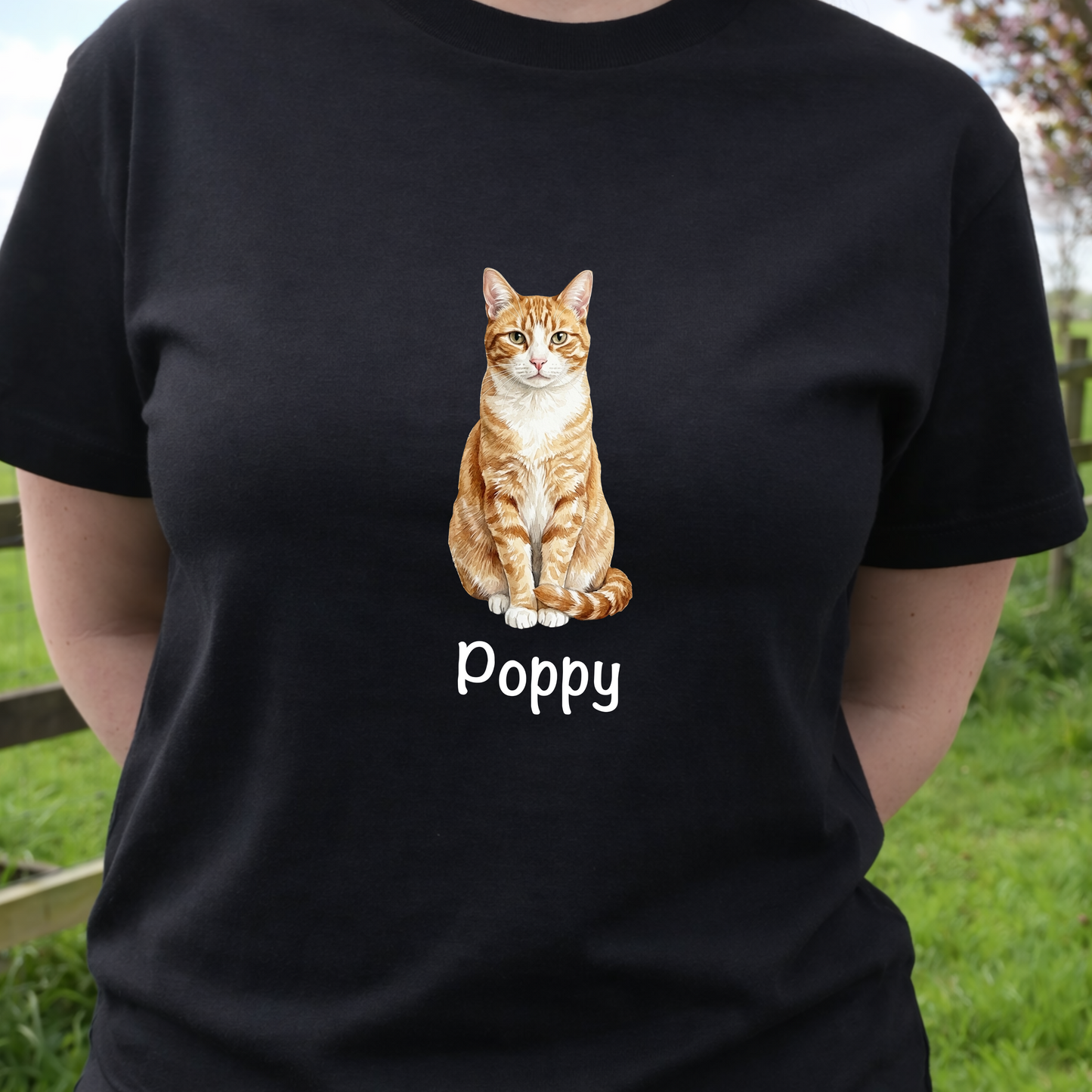 Person wearing a personalised cat black t-shirt with a watercolour ginger cat illustration and 'Poppy' text, standing outdoors.