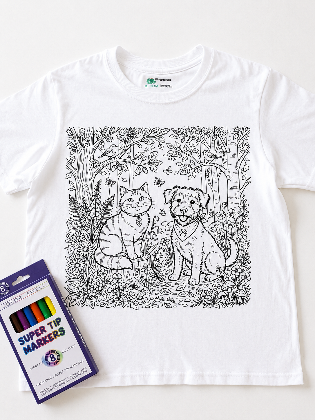 A white short-sleeve T-shirt laid flat on a white background, featuring a black line-art illustration of a cat and a dog in a forest scene on the front. A package of Super Tip Markers is placed at the lower left as a styling prop.