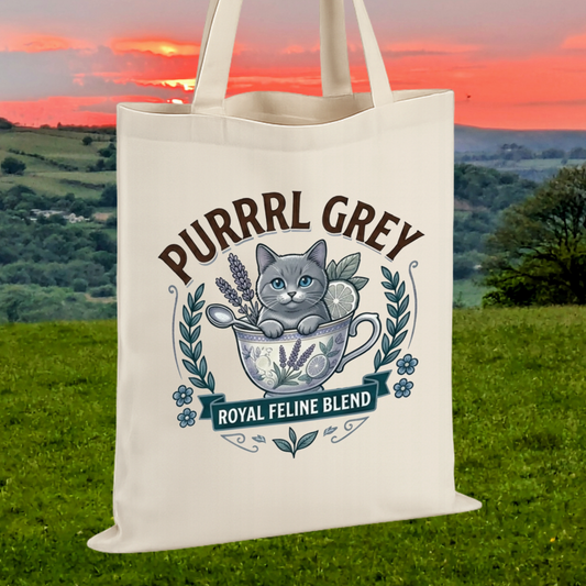 An off-white canvas tote bag with a printed graphic of a grey cat in a teacup with lavender and flowers, under the text 'PURRRL GREY' and 'ROYAL FELINE BLEND', shown against a scenic background of a grassy field and hills at sunset.