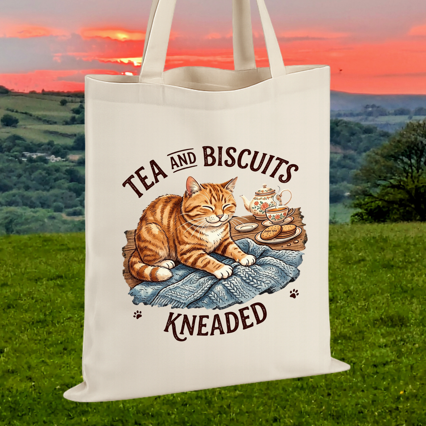 An off-white canvas tote bag with a printed illustration of an orange tabby cat lounging on a blue blanket next to a teapot and biscuits, with the text 'TEA AND BISCUITS KNEADED' in dark brown. 